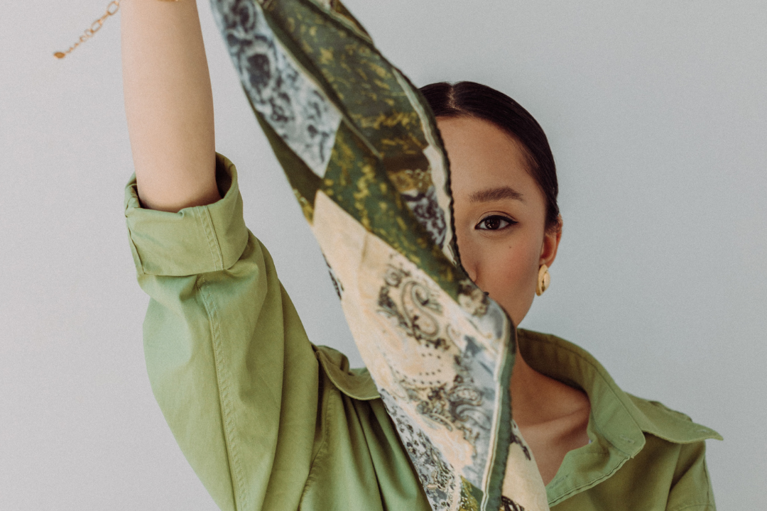 Woman wearing a green jacket and holding a patterned scarf against a plain background